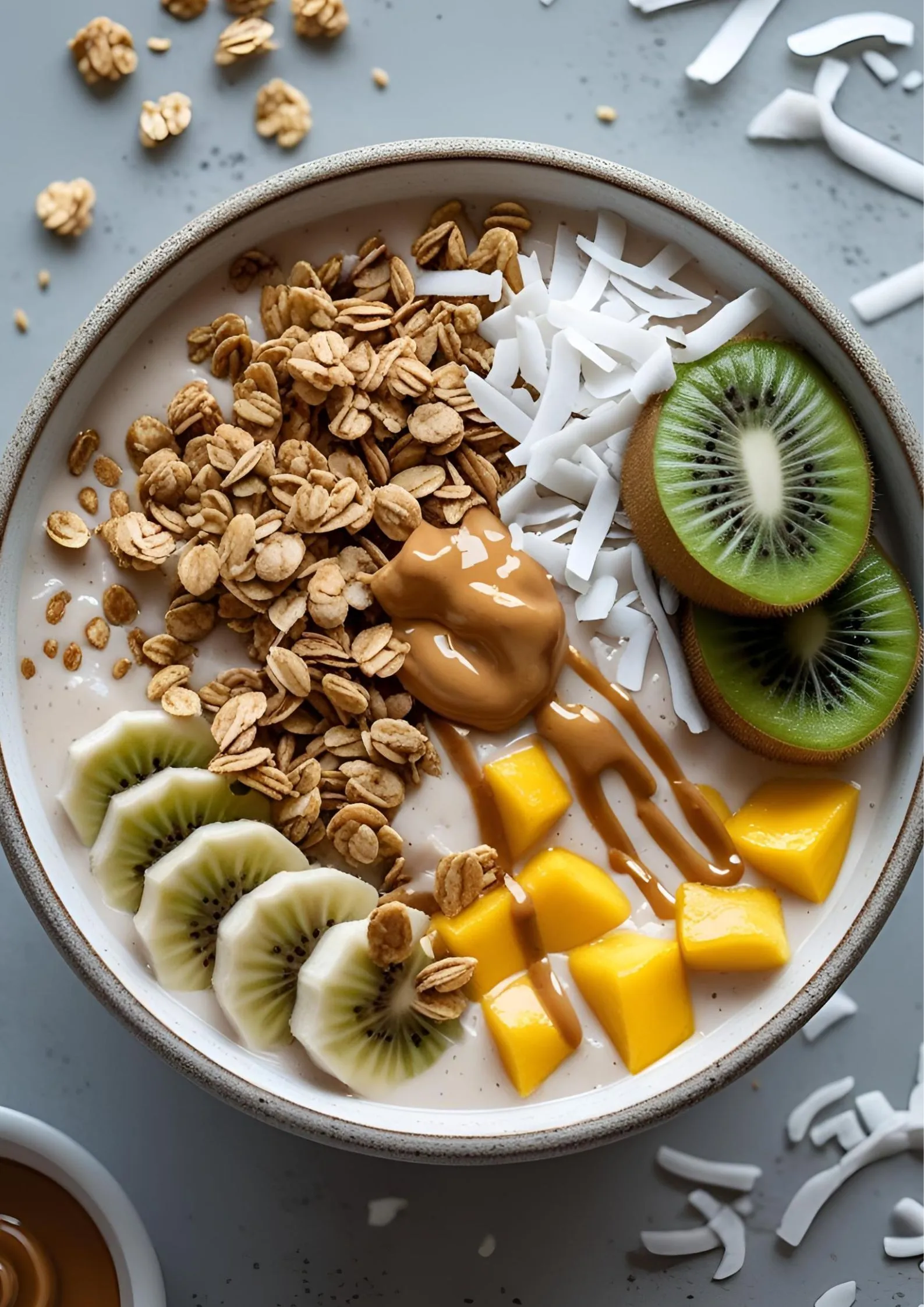 A vibrant smoothie bowl with blended mango and banana, topped with granola, sliced kiwi, shredded coconut, and almond butter drizzle.