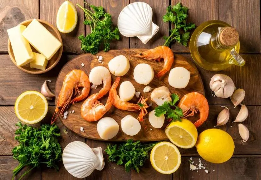 Fresh ingredients for garlic butter seafood recipe including shrimp, scallops, lobster, garlic, parsley, lemon, butter, and olive oil on wooden board.