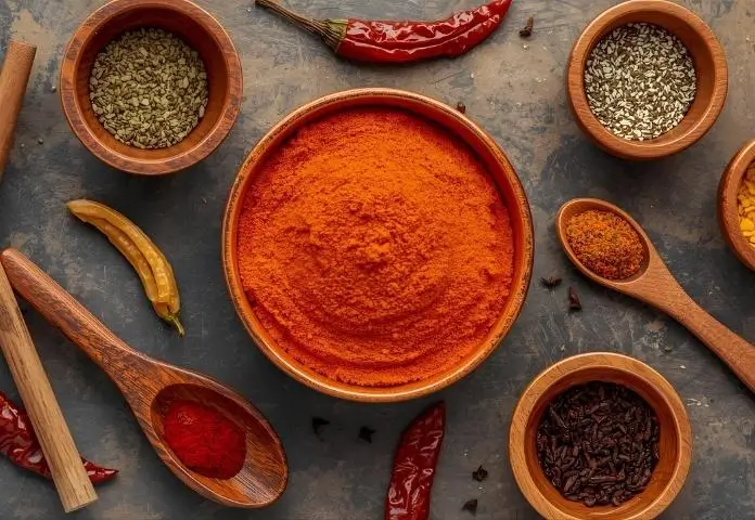 A bowl of colorful birria seasoning powder is surrounded by cumin, oregano, cloves, dried chilies, and wooden spoons.