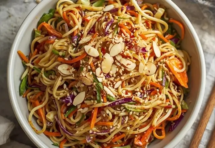 A bowl of easy ramen noodle salad with toasted ramen noodles, carrots, green and purple cabbage, and almonds on a wooden table.