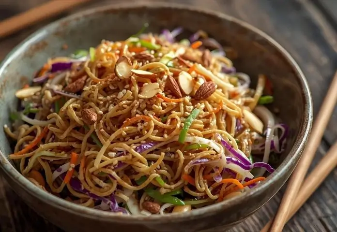 A close-up of a crunchy ramen noodle salad with toasted ramen noodles, shredded cabbage, carrots, almonds, and sesame seeds in a rustic bowl.