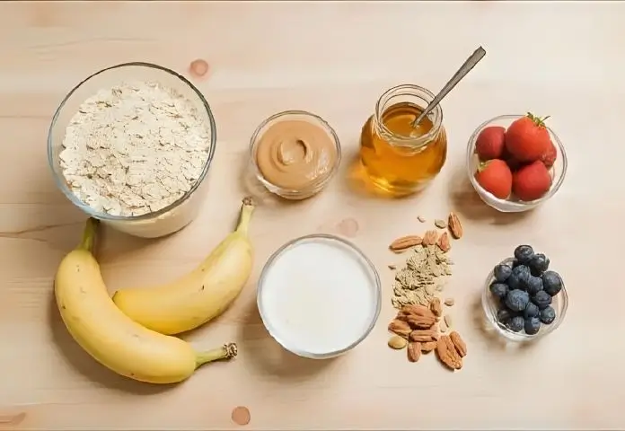 A flat lay of the ingredients for protein oatmeal on a wooden background shows rolled oats, a scoop of protein powder, a banana, a jar of nut butter, fresh berries, almonds, chia seeds, and a jar of honey.