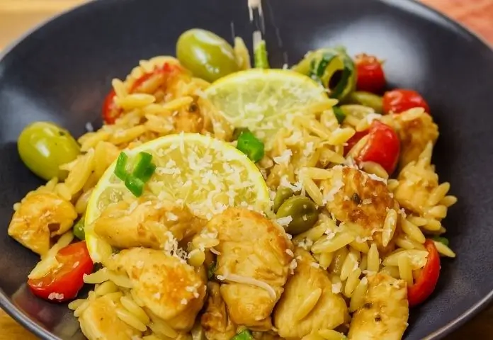A rustic skillet filled with Mediterranean Chicken Orzo featuring tender chicken pieces, halved cherry tomatoes, green and black olives, fresh parsley, and lemon slices, all set on a wooden table.