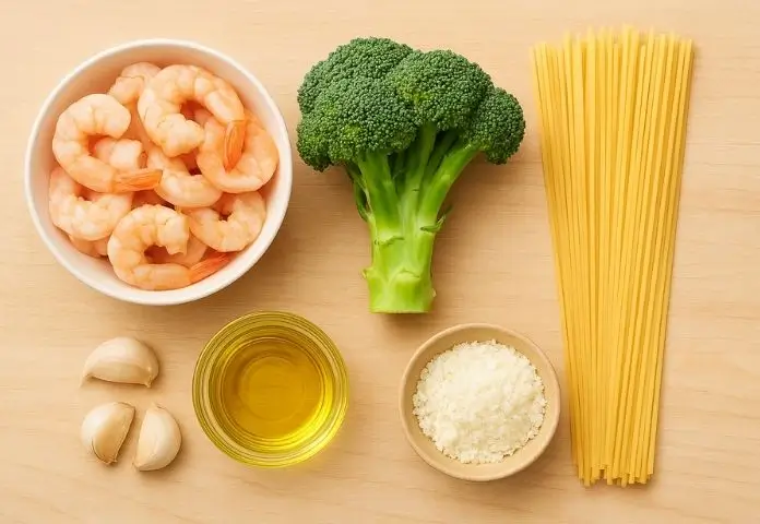 Fresh peeled shrimp, green broccoli florets, uncooked pasta, garlic cloves, lemon slices, butter, olive oil, and basil leaves arranged on a wooden surface.